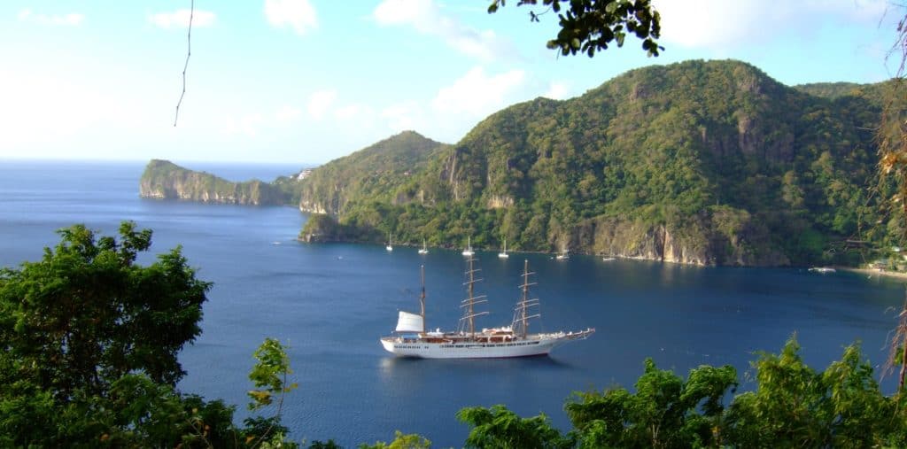 A tall ship with white sails anchors in a blue bay surrounded by steep, forested islands.