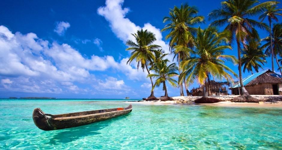 A wooden boat floats in turquoise water near a tropical beach with palm trees and thatched.