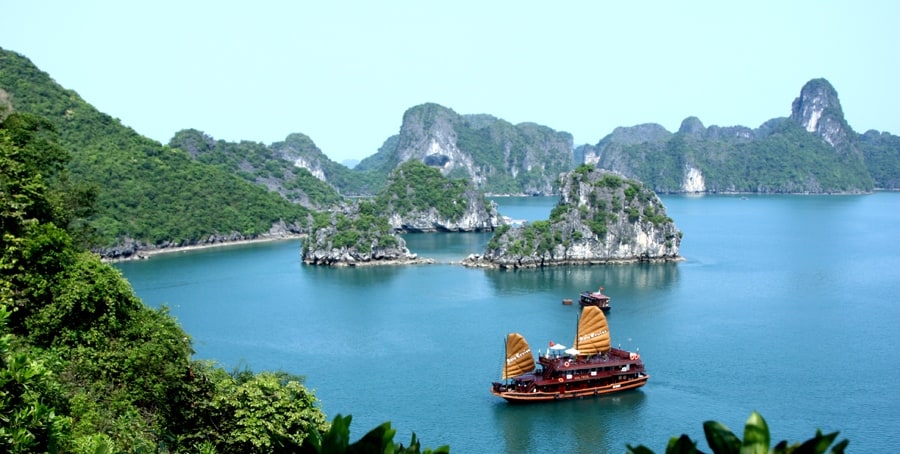 Traditional wooden junk boat with golden sails on turquoise water surrounded by limestone karst.
