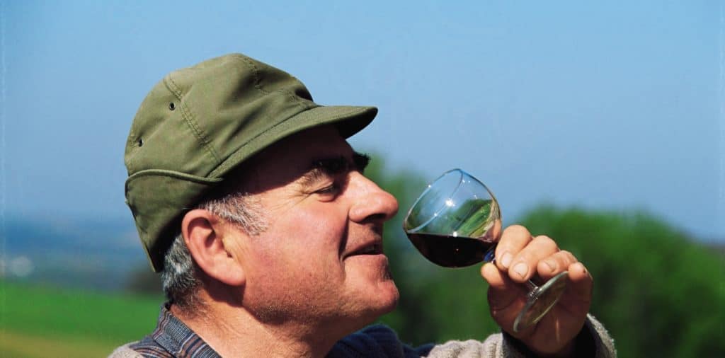 Man in a khaki cap holding a glass of red wine outdoors.
