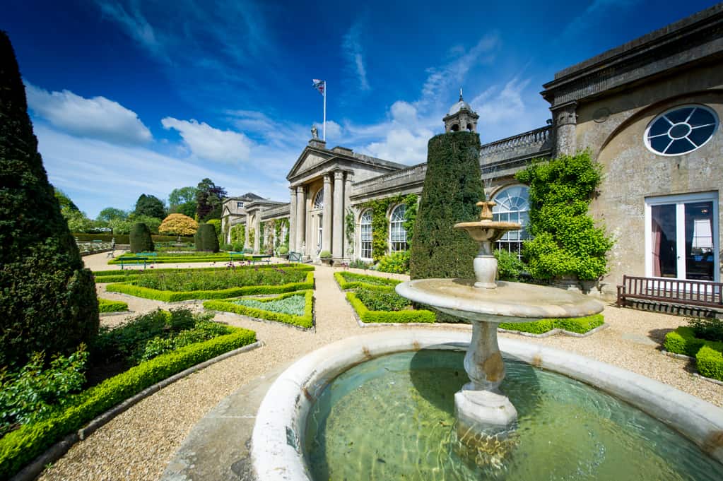 A formal garden with a circular fountain in the foreground and a classical stone building.