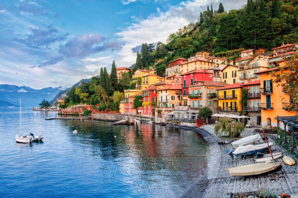 Colorful waterfront village with boats on a blue lake surrounded by forested mountains.