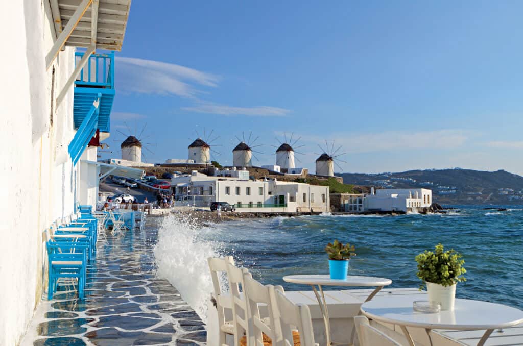Waterfront café with blue chairs overlooking windmills and sea in Mykonos, Greece.