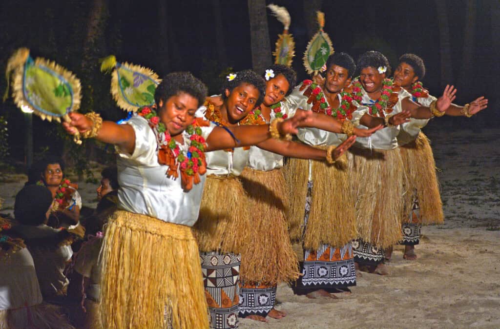 A line of dancers in traditional Pacific Islander attire perform with fans and grass skirts.