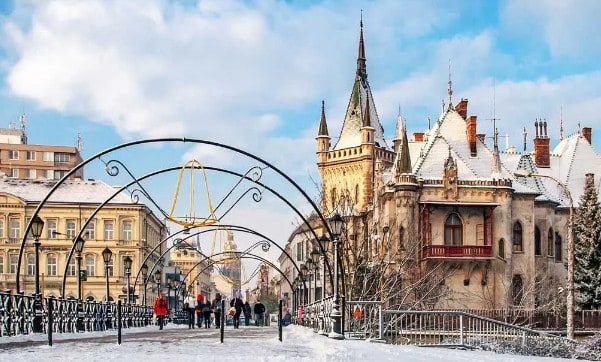 A Gothic Revival building with a spire stands beside an ornate metal arch in a public square.