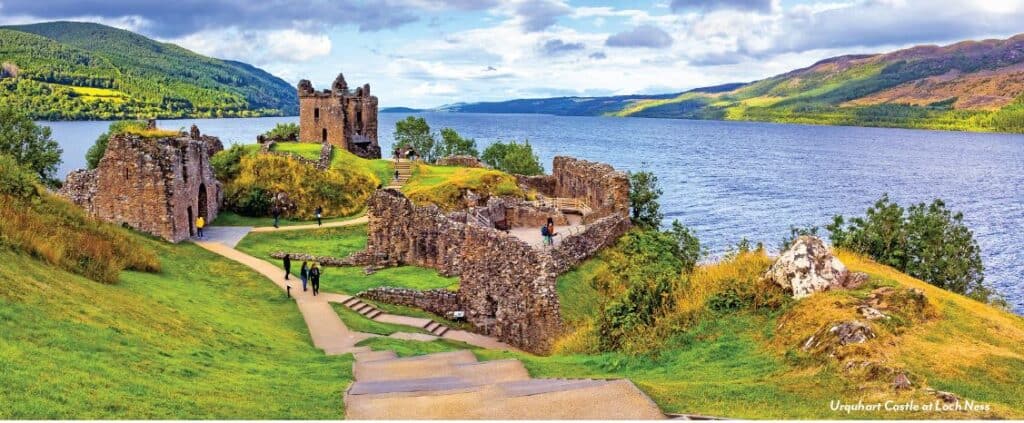 Urquhart Castle ruins overlook Loch Ness with green hills and mountains in the Scottish Highlands.
