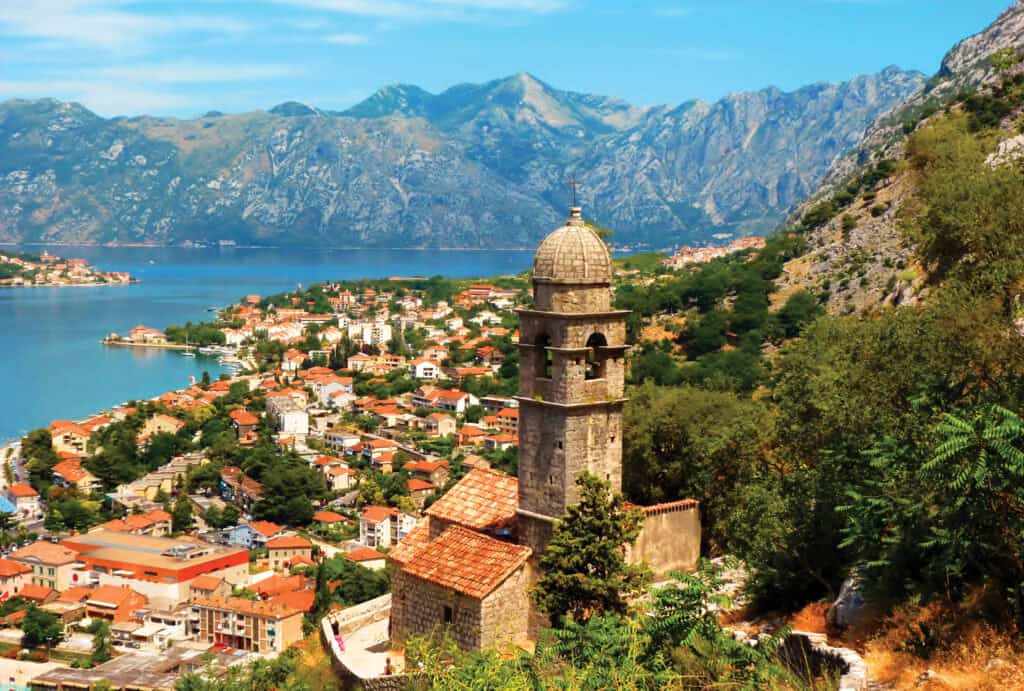 Stone minaret tower overlooks a coastal Mediterranean town nestled between mountains and blue water.