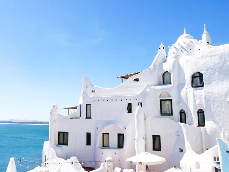 White-washed building with distinctive architecture overlooking turquoise sea under clear blue sky.